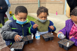 Kids wearing winter jackets and masks plant seeds in cartons while standing at a table outside