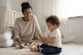 smiling woman with her hair pulled up sits on the floor playing with her baby, who is sitting up and holding a toy.